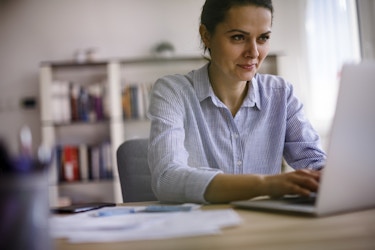  A young woman in a button-up shirt works on a laptop. Behind her, out of focus, is a book-lined set of shelves, and there are papers and pens on the desk beside her. 